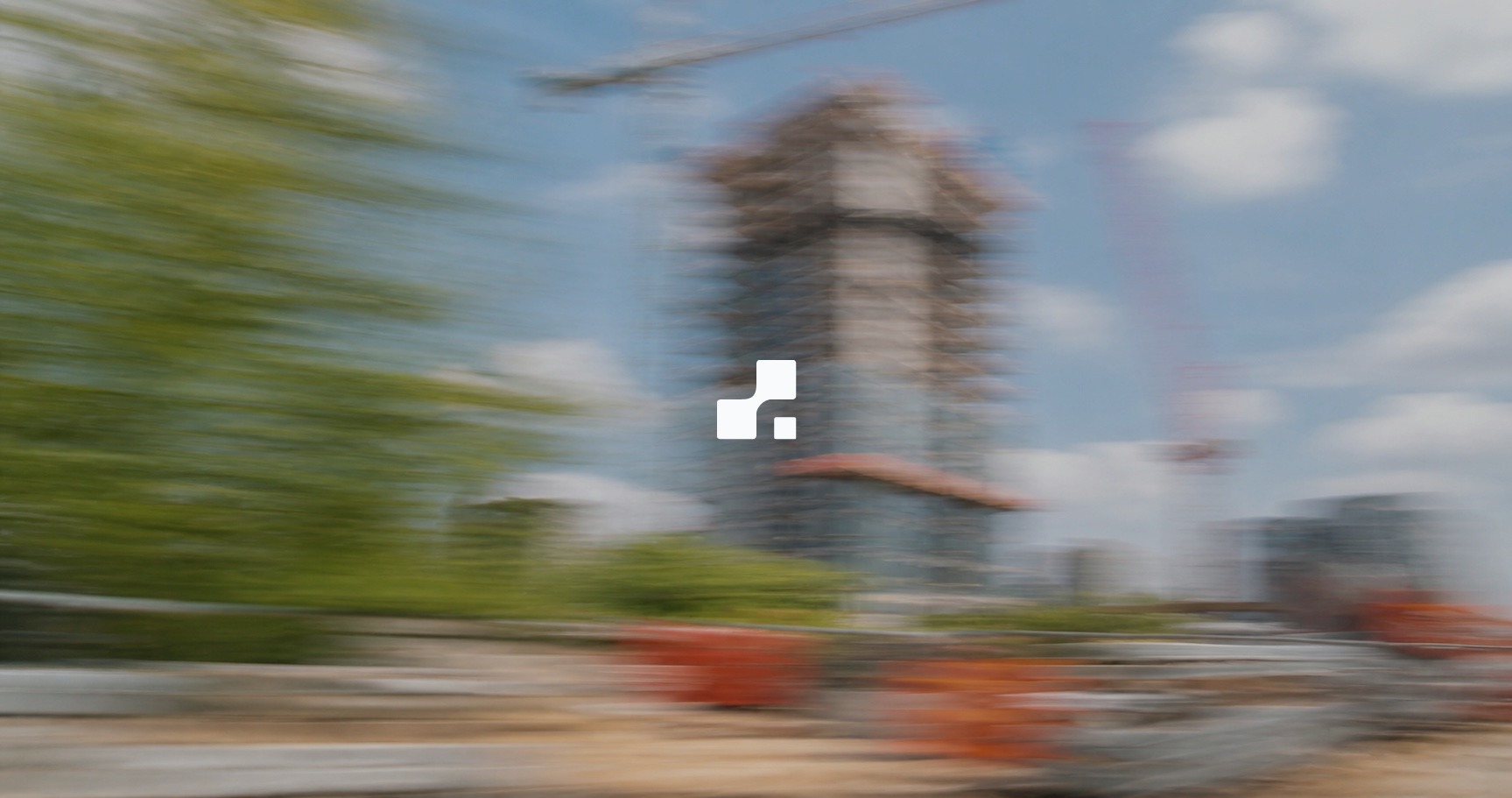 Vue estivale floutée par le mouvement d’une tour en construction, avec des grues et des arbres verts, sur laquelle figure le symbole Aplon