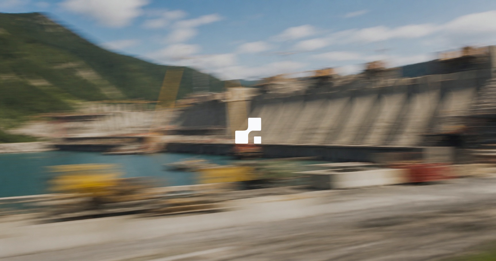 Vue estivale floutée par le mouvement d’un barrage en construction dans des montagnes verdoyantes, sur laquelle figure le symbole Aplon