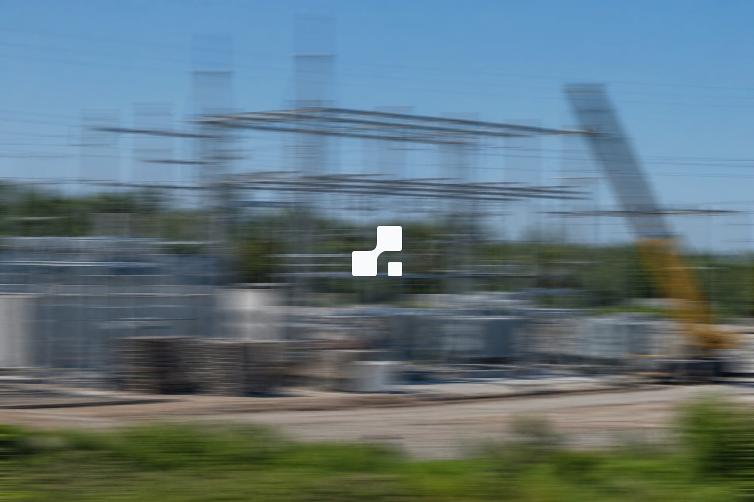 Vue estivale entièrement floutée par un mouvement latéral d’un poste électrique en construction, avec des structures d’acier, une grue et de la végétation, sur laquelle figure le symbole Aplon